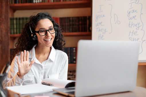 Distance Education. Positive lady wearing glasses and wireless headest at virtual meeting, sitting at desk, having video call on laptop, waving to webcam. Woman studying or teaching online at home